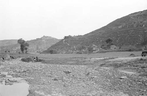 Group of dwellings at foot of hill, river in front of them. Road with car on it