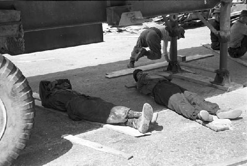 Men laying on wooden planks under truck avoiding the sun