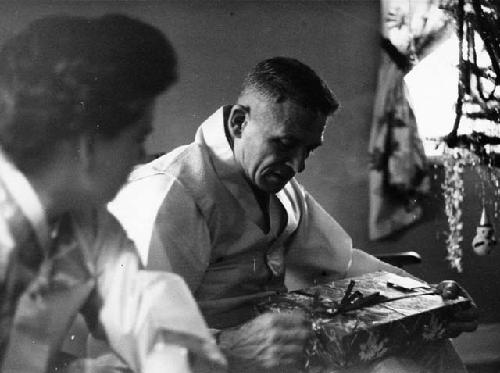 General Whitcomb opening present in wrapping paper, sitting next to Lieutenant M