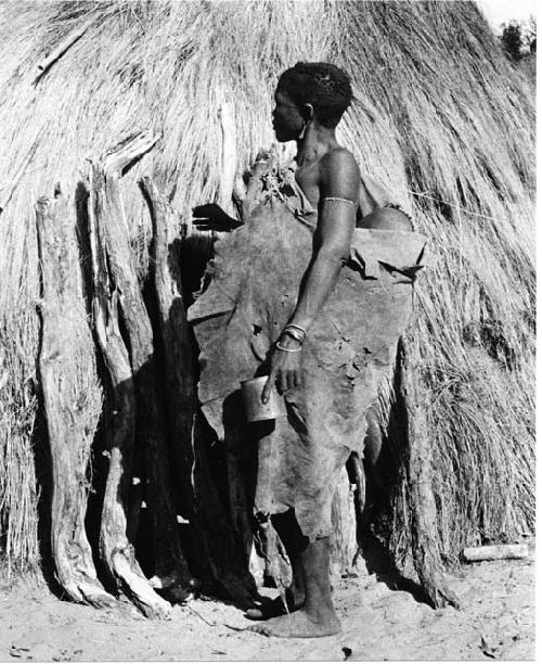 Woman with a kaross filled with something standing in front of a hut