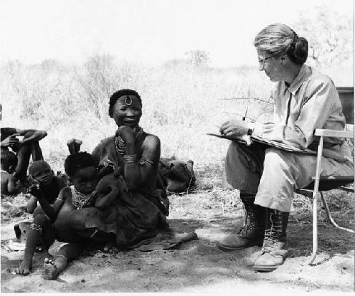 Lorna Marshall taking notes, !Ungka Norna seated between !U's legs, and other unidentified people