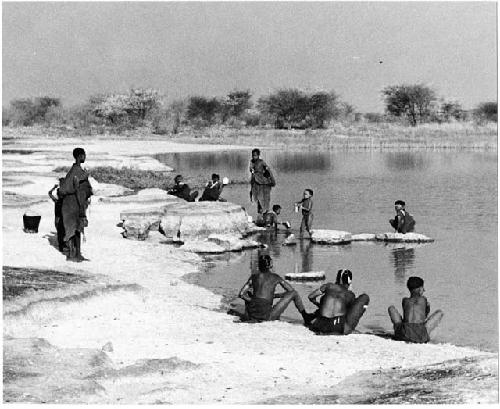 Group of people beside the water; some women are bathing, and others are filling ostrich egg shells with water