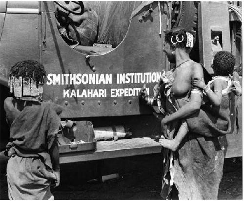 N!ai and a woman standing in front of one of the expedition's GMC trucks with "Smithsonian Institution Kalahari Expedition" showing