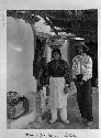 Husband and wife stand outside dwelling; woman carries bowl on head