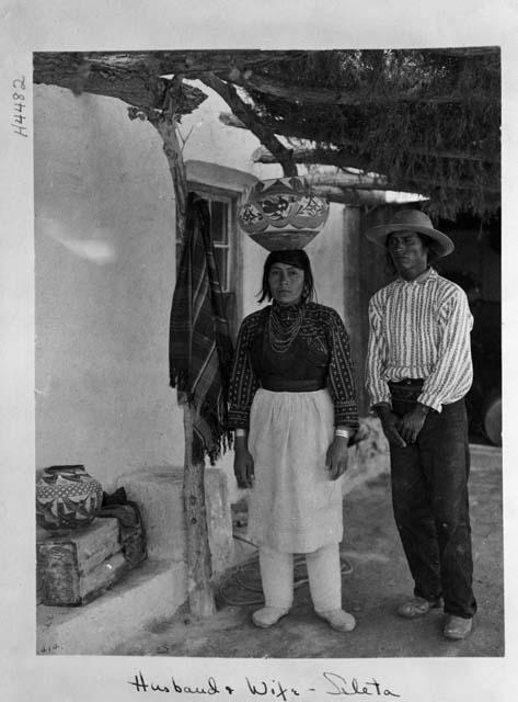 Husband and wife stand outside dwelling; woman carries bowl on head
