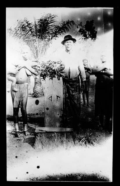 Four males standing; three shirtless boys with folded arms (one holding small child), one man wearing shirt and hat