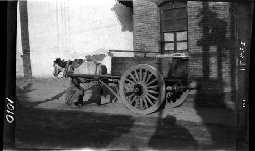 Man squatting next to horse-drawn cart; brick building in back