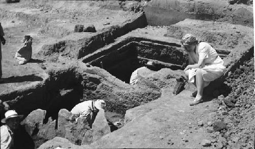 Woman seated at excavation site, man working in pit
