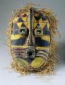 Carved wood mask with black, yellow, and red painted decorations and raffia trim