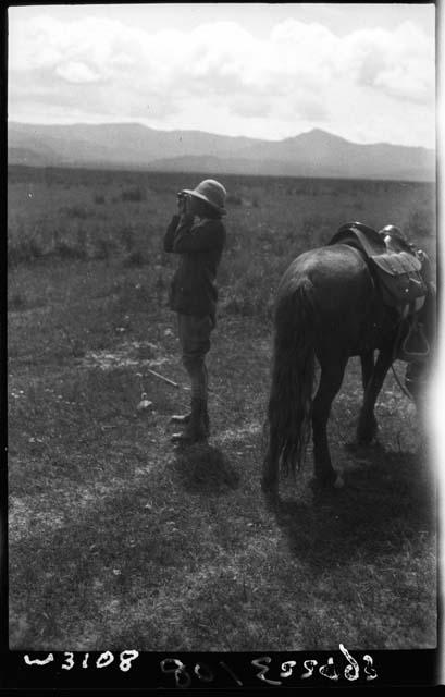 Woman looking through binoculars stands next to horse