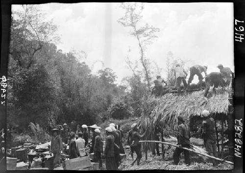 People gather around workers building hut, crates on ground