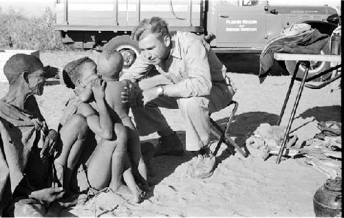 Tsekue and another person sitting, with N!whakwe standing next to them, talking with Daniel Blitz, expedition truck in the background