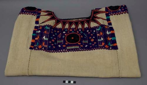 Huipil, six panels sewn together, light beige ground with black fabric appliqué and purple, green, and orange brocade along chest, neckline and shoulders. Light beige ground is stained and has darkened over time.