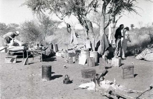 Cooking fire in the expedition camp, with Claude McIntyre with his belongings, Charles Koch and Laurence Marshall walking in the background