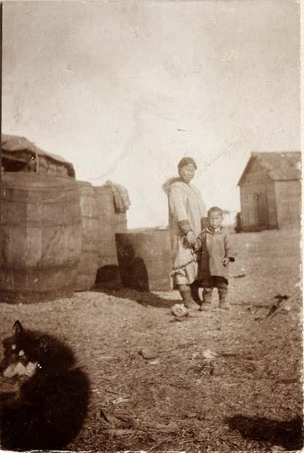 Inuit woman and child standing in front of barrels and wood frame buildings