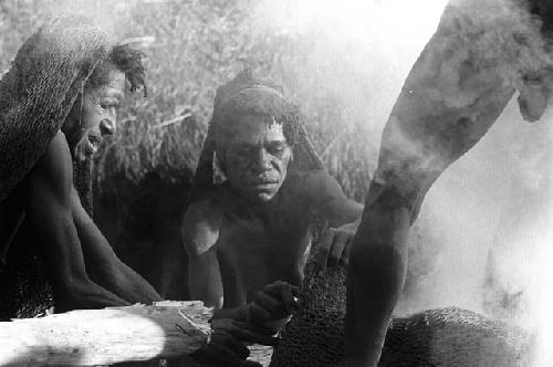 Woman put on the fire;  man stands on the log and helps;  2 women in the background helping also