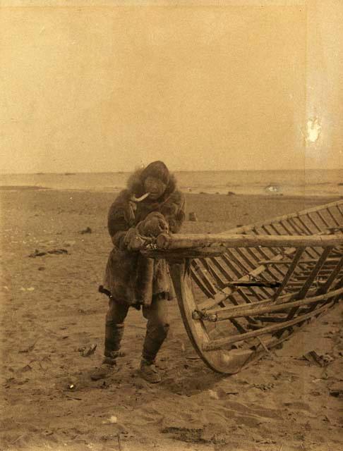 Inuit man leaning against the frame of an umisk and smoking a pipe ...