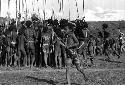 Samuel Putnam negatives, New Guinea; little boy running with a toy bow and arrow; behind him is a group singing; of men and boys
