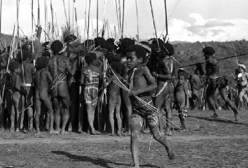 Samuel Putnam negatives, New Guinea; little boy running with a toy bow and arrow; behind him is a group singing; of men and boys
