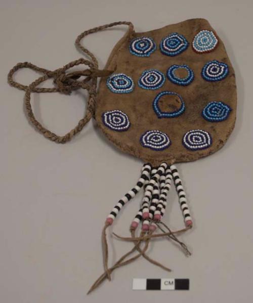 Medium leather bag beaded on one side with light blue, dark blue and white circular motifs; and one light blue, white and red circular motif; 7 pendent strings of black, white and pink glass beads at base of bag; long twisted leather strap