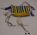 Medium leather bag with attached beaded panel. Beaded panel has dark blue and yellow vertical bands, horizontal bands of yellow and black chevrons, blue and orange; white edging. Pendant strings of large white beads, dark blue oval beads, one red oval bead, one mother of pearl button; long twisted leather strap