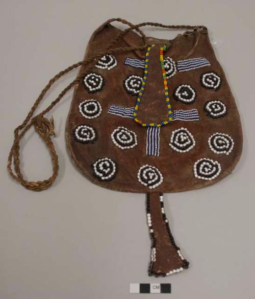 Large leather bag beaded on one side with black and white circular motifs, blue and white striped motifs; leather element at bag opening edged with green, yellow, blue and orange beads; dangling leather element at base of bag edged with black and white beads; long twisted leather strap