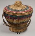 Basket with dyed pink, green, purple and dark blue pattern; lid with woven knob; leather woven into basket; leather strap