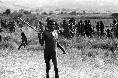 Man dancing with a long object (spear?), women's group beyond him