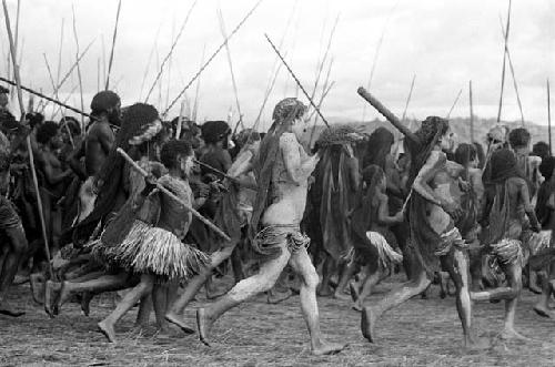 Large group of men and women dancing; spears show over heads; woman in foreground covered in clay