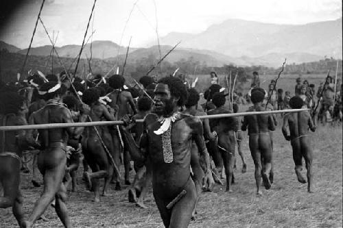 Men dancing at an Etai, women's group beyond them; man in the foreground passes with spear leaning horizontally on shoulder