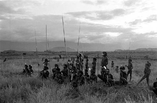 Men and boys watch an Etai, spears stuck vertically in the ground around them