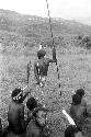 Men sitting and watching the Etai; man standing in front with long spear and clay on his legs