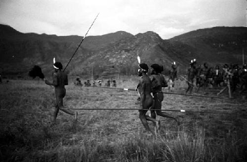 Men dancing, spears in hand -- others dancing in background; hills beyond