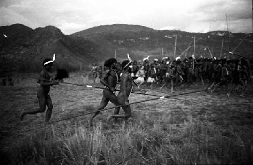 Men dance, spears in hand -- large group of dancers beyond them