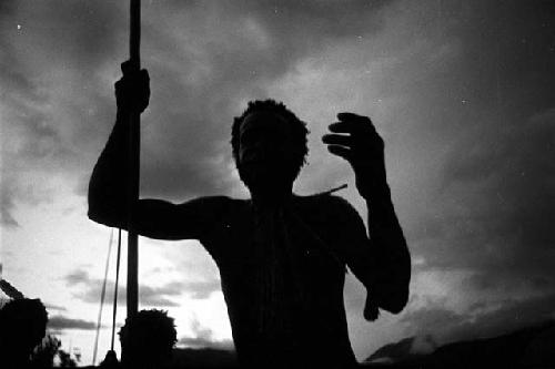Silhouetted man leaning on his spear, gesturing with a hand