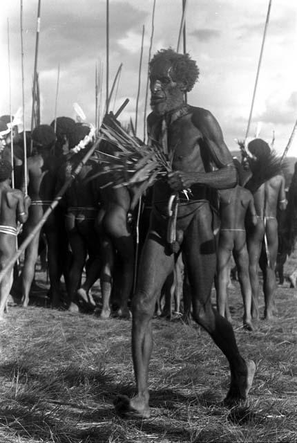 Dancing man at an Etai with spear and leaves in his hands; others beyond