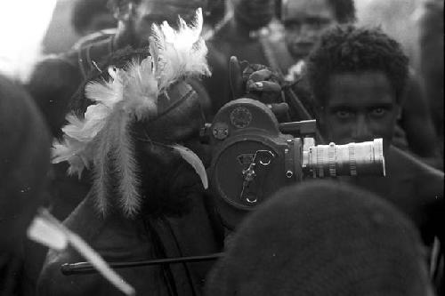 Man with feather headdress looks through Filmo video camera