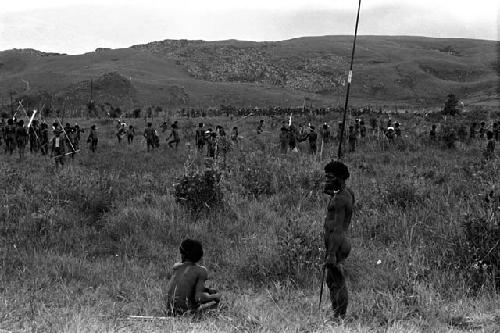 Two men waiting in the foreground as a large group beyond them comes back from battle on the Tokolik; hills and war beyond