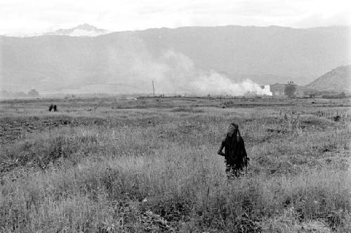 Women working in a field; a fire burns in the distance