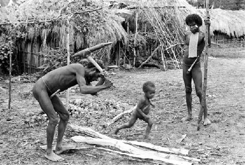 Loliluk splits wood with a stone axe; Natolek runs between him and Wali who watches