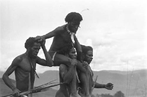 Four men walking, one carrying another man on his shoulders