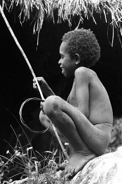 Yegé Abu sitting on a rock with a hoop and spear