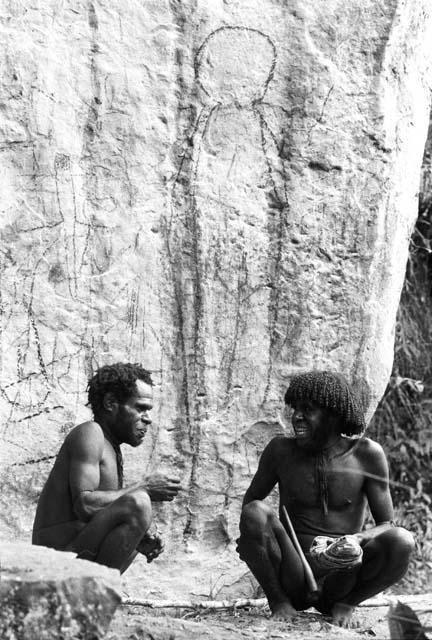 Two men beneath a rock shelter with drawings behind them