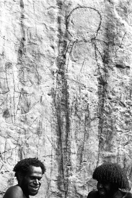 Two men beneath a rock shelter with drawings behind them