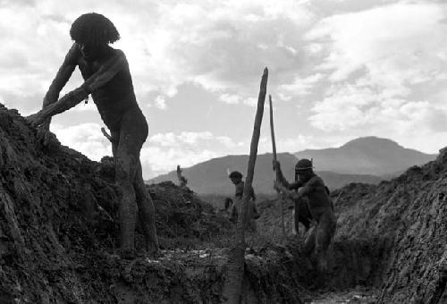 Man smoothing the sides of the ditch behind people working digging at the trash