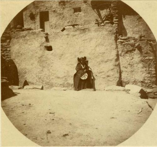 Hopi woman standing and holding a basket in front of the pueblo