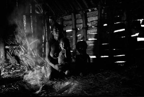 A woman and 2 children sitting by a fire in the hunu at Lokoparek