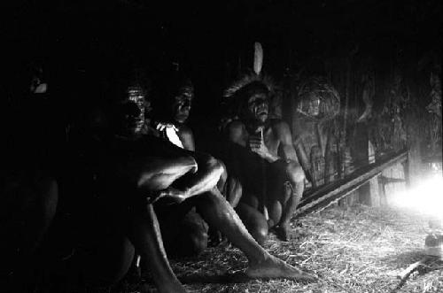 Men seated around the wall of the honai at Lokoparek; Coleman lamp flares on the right