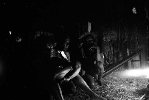 Men seated around the wall of the honai at Lokoparek; Coleman lamp flares on the right