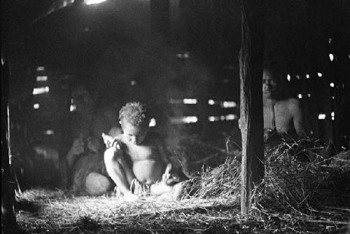 A little girl sits by a fire in the hunu at Wuperainma II; 2 others behind her